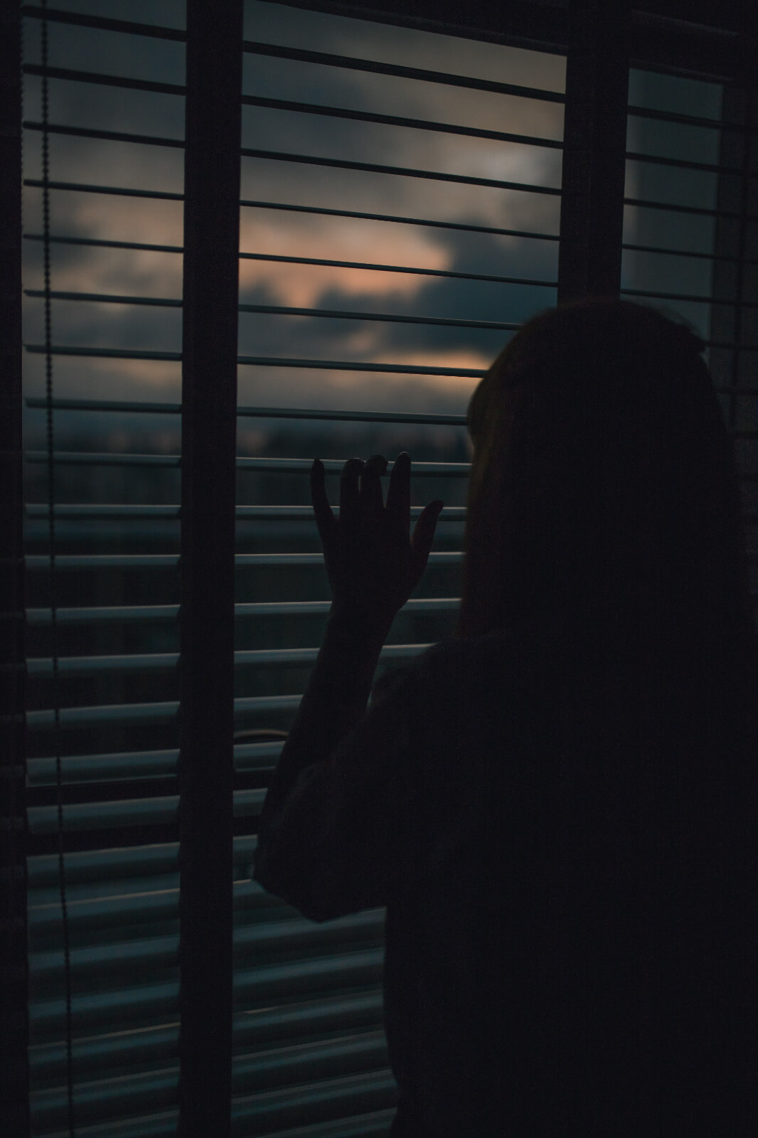 Photo of a woman peeking out of her blinds at sunset