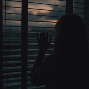 Photo of a woman peeking out of her blinds at sunset